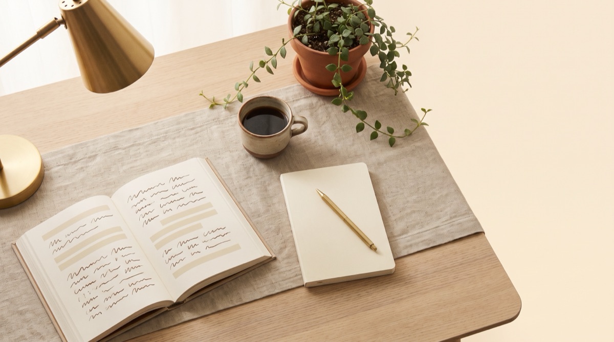 A light academia study desk from above: pale oak, a linen runner, an open book, a cream notebook with a brass pen, a ceramic cup of coffee, a trailing plant and a brass lamp.